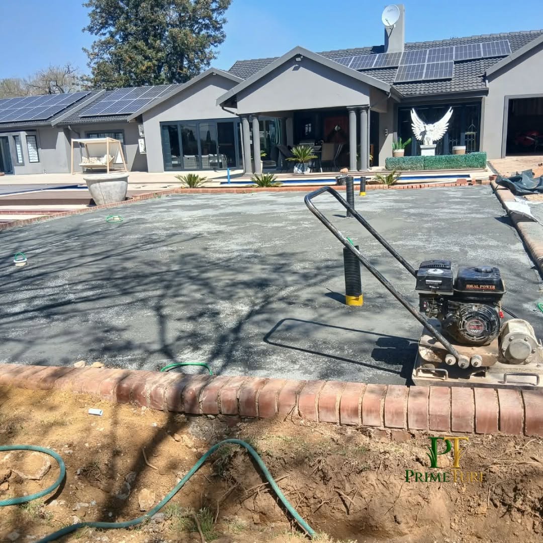 Compacted stone base being prepared with a plate compactor inside a brick edge, house in the background