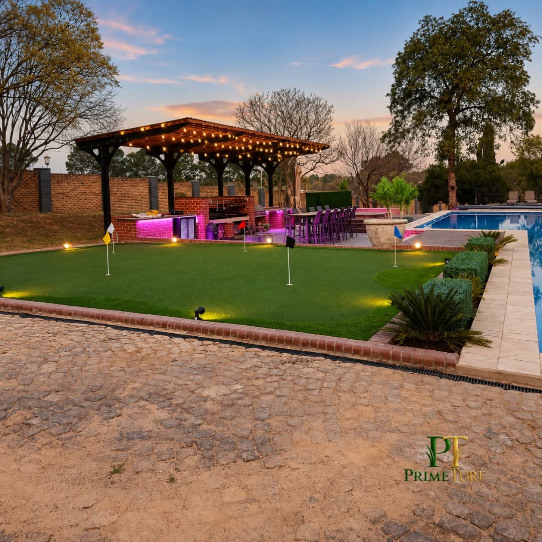 The same putting green at dusk with garden lighting on, pergola bar and pool alongside